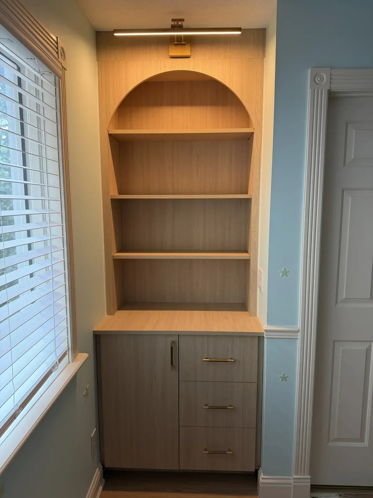 Custom built-in with an arched bookcase, brass light, and toy storage base cabinet between a window and door in a Calgary kids' room